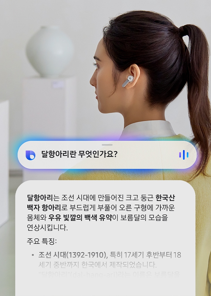 한쪽 귀에 화이트 색상 갤럭시 버즈4 프로를 착용한 여성의 뒷모습이 있습니다. 옆에는 AI에게 달항아리란 무엇인가요?라고 묻는 말풍선이 있습니다. 말풍선 아래에는 달항아리는 달항아리는 조선 시대에 만들어진 크고 둥근 한국산 백자 항아리로 부드럽게 부풀어 오른 구형에 가까운 몸체와 우유 빛깔의 백색 유약이 보름달의 모습을 연상시킵니다. 라는 답변이 담긴 다른 말풍선이 있습니다. 그 아래에는 달항아리의 주요 특징들이 나열되어 있습니다.