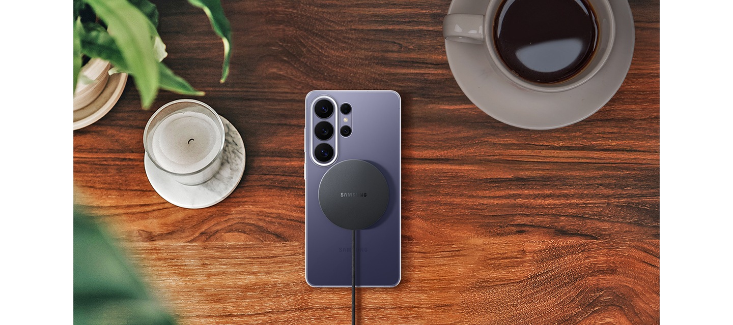 나뭇결 지면 위로 클리어 마그넷 케이스를 장착한 코발트 바이올렛 색상의 갤럭시 S26 울트라가 놓여져 있습니다. 그 뒤로 마그넷 무선 충전기가 정렬되어 있고 이를 통해 간편한 충전을 강조하고 있습니다.