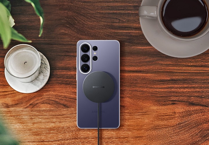 나뭇결 지면 위로 클리어 마그넷 케이스를 장착한 코발트 바이올렛 색상의 갤럭시 S26 울트라가 놓여져 있습니다. 그 뒤로 마그넷 무선 충전기가 정렬되어 있고 이를 통해 간편한 충전을 강조하고 있습니다.