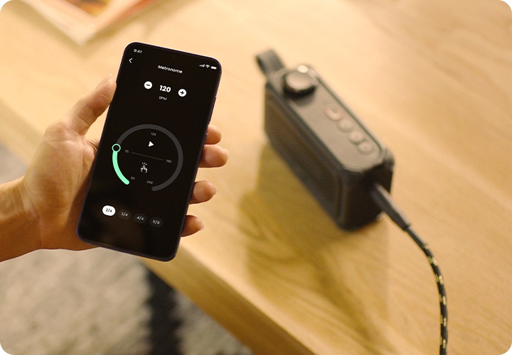 테이블 위에 BANDBOX SOLO가 놓여 있으며, 사람 손에는 휴대폰이 들려 있습니다. 휴대폰 화면에는 BANDBOX SOLO와 연결된 앱 화면이 켜져 있습니다.