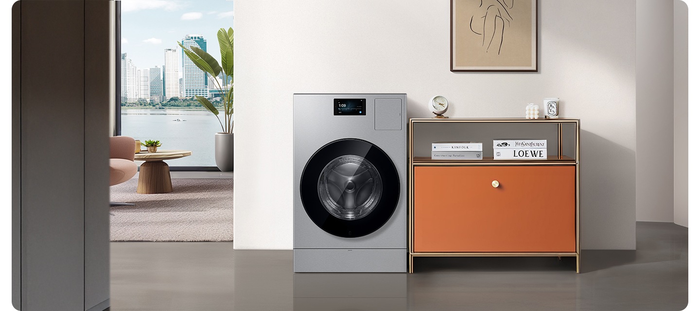 화이트톤 벽면에 AI 콤보가 설치되어 있습니다. 제품 우측에는 오렌지색 수납장이 놓여있고 각종 오브제가 올려져 있습니다. 윗쪽 벽면에는 그림액자가 설치되어 있습니다. 제품 좌측으로는 의자와 테이블이 놓여있고 통창으로 외부 건물과 강이 보이는 연출된 인테리어 이미지 입니다.