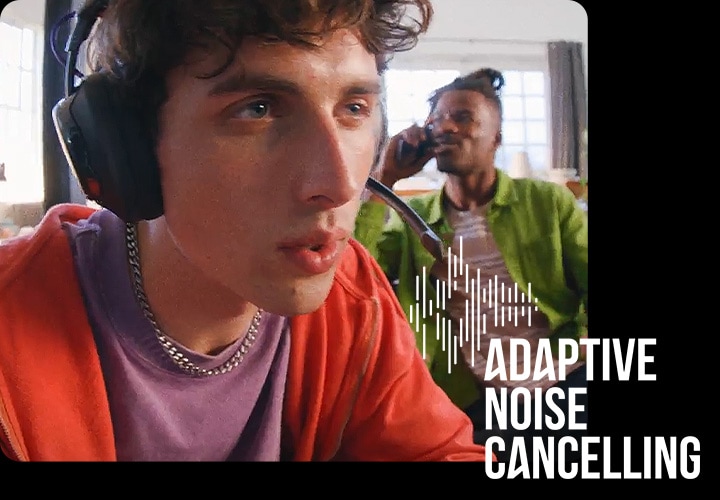 헤드폰을 착용한 남성이 게임을 하고 있으며, 그 뒤로는 통화를 하고 있는 남성이 있습니다.