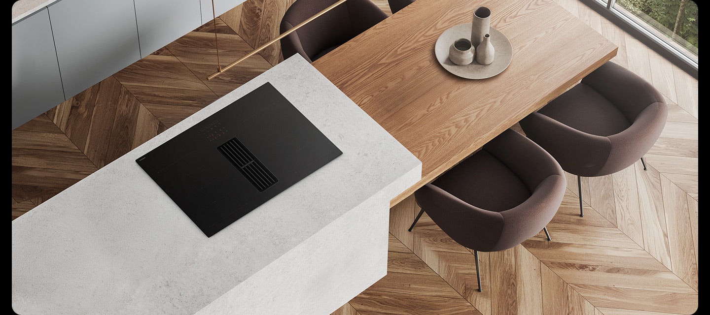 헤링본 마루가 깔린 바닥과 화이트 톤의 주방을 위에서 내려본 각도를 배경으로 이미지 왼쪽 부분에는 화이트 색상의 상판과 설치된 Infinite Line 후드일체형 인덕션이 노출되고 있고 인덕션 오른쪽 부분에는 상판과 연결되어 있고 우드로 제작된 테이블이 있습니다. 테이블 주위에는 진한 브라운 색상에 1인용 의자 4개가 놓여있습니다.