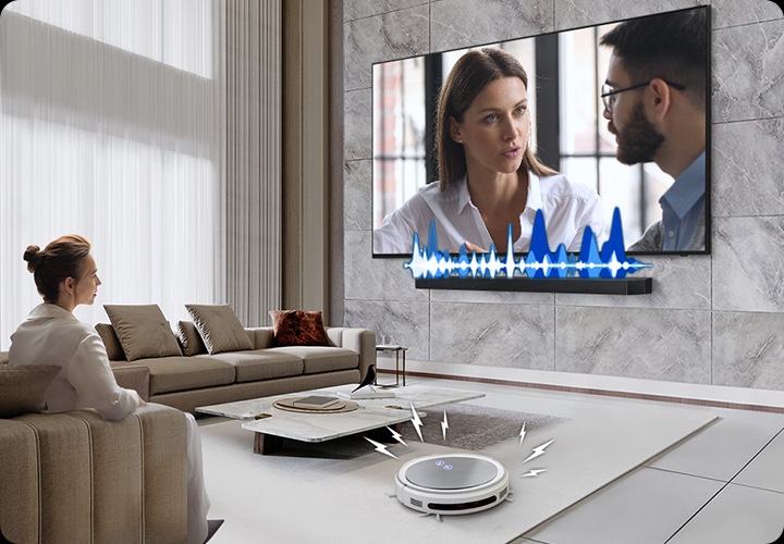 거실에서 여성이 TV를 보고 있습니다. TV 화면에는 드라마에 한 장면이 나오고 있습니다. TV 하단에는 사운드바가 설치되어 있고 TV와 사운드바 사이에는 음향효과가 있습니다. 거실 바닥에는 무선 로봇 청소기가 움직이고 있습니다.