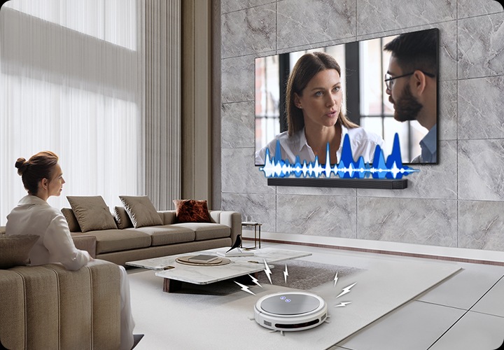 거실에서 여성이 TV를 보고 있습니다. TV 화면에는 드라마에 한 장면이 나오고 있습니다. TV 하단에는 사운드바가 설치되어 있고 TV와 사운드바 사이에는 음향효과가 있습니다. 거실 바닥에는 무선 로봇 청소기가 움직이고 있습니다.