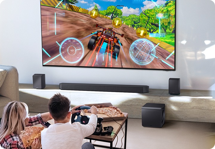 벽에 TV가 설치되어 있습니다. TV 아래에는 사운드바와 리어 스피커가 놓인 장식장이 있으며, 장식장 앞에는 서브우퍼가 놓여 있습니다. TV 앞에는 남성과 여성이 거실에 앉아서 게임을 즐기고 있습니다.