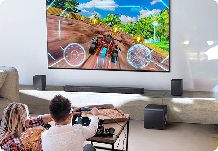 벽에 TV가 설치되어 있습니다. TV 아래에는 사운드바와 리어 스피커가 놓인 장식장이 있으며, 장식장 앞에는 서브우퍼가 놓여 있습니다. TV 앞에는 남성과 여성이 거실에 앉아서 게임을 즐기고 있습니다.