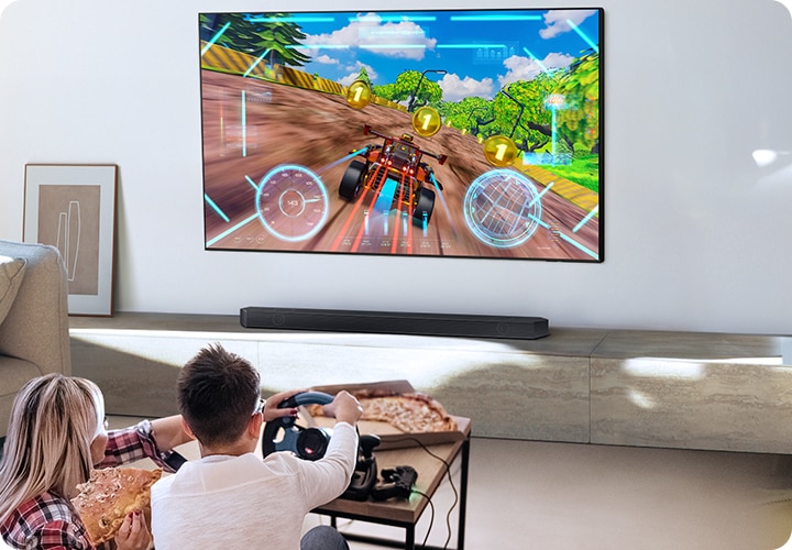 벽에 TV가 설치되어 있습니다. TV 아래에는 사운드바가 놓인 장식장이 있습니다. TV 앞에는 남성과 여성이 거실에 앉아서 게임을 즐기고 있습니다.