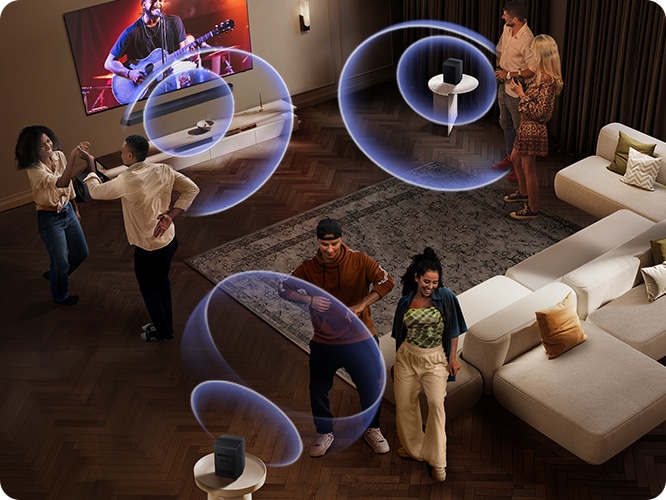 거실 벽에 TV와 사운드바가 설치되어 있습니다. TV 화면에는 공연장에서 사람들이 공연을 즐기고 있습니다. 사운드바와 소파 뒤에 연결된 리어 스피커에서 음향효과가 나오고 있습니다. 사람들이 거실 중앙에서 춤을 추고 있습니다.