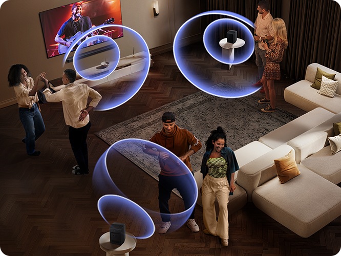 거실 벽에 TV와 사운드바가 설치되어 있습니다. TV 화면에는 공연장에서 사람들이 공연을 즐기고 있습니다. 사운드바와 소파 뒤에 연결된 리어 스피커에서 음향효과가 나오고 있습니다. 사람들이 거실 중앙에서 춤을 추고 있습니다.