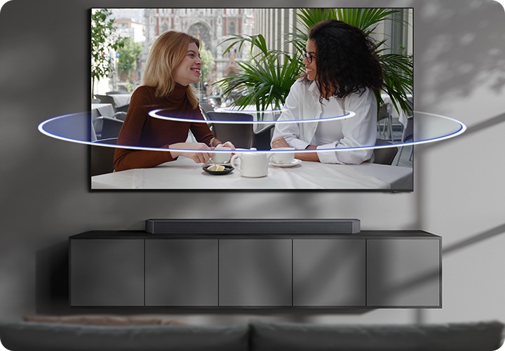 벽에 TV가 설치되어 있으며, TV 아래에는 사운드바가 놓인 장식장이 있습니다. TV 화면에는 여성 두명이 티타임을 가지고 있습니다. TV 화면에서 음향효과가 나오고 있습니다.