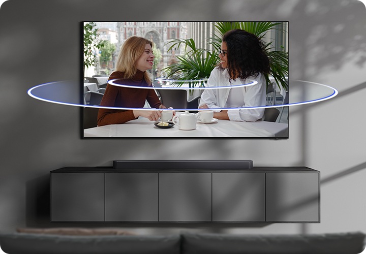 벽에 TV가 설치되어 있으며, TV 아래에는 사운드바가 놓인 장식장이 있습니다. TV 화면에는 여성 두명이 티타임을 가지고 있습니다. TV 화면에서 음향효과가 나오고 있습니다.