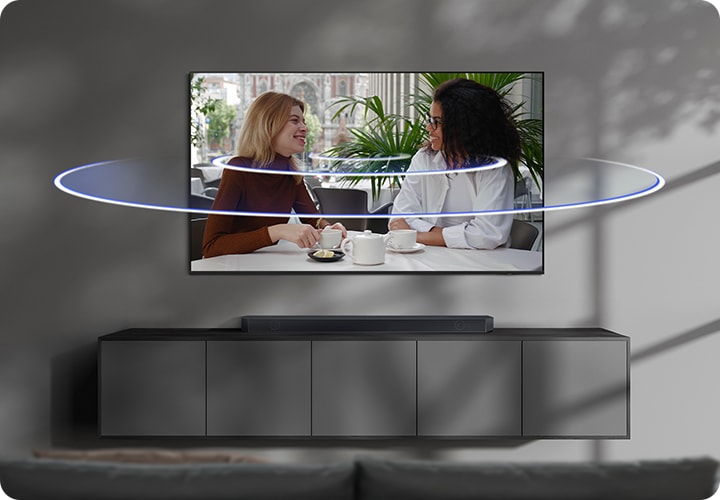 벽에 TV가 설치되어 있으며, TV 아래에는 사운드바가 놓인 장식장이 있습니다. TV 화면에는 여성 두명이 티타임을 가지고 있습니다. TV 화면에서 음향효과가 나오고 있습니다.