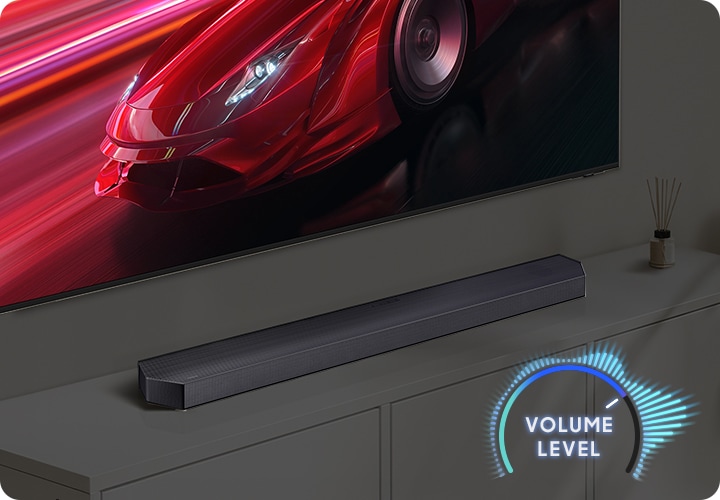 벽에 TV가 설치되어 있으며, TV 아래에는 사운드바가 놓인 장식장이 있습니다. TV 화면에는 붉은색 자동차가 송출됩니다. 이미지 오른쪽 하단에는 볼륨 레벨이 표시되어 있습니다.