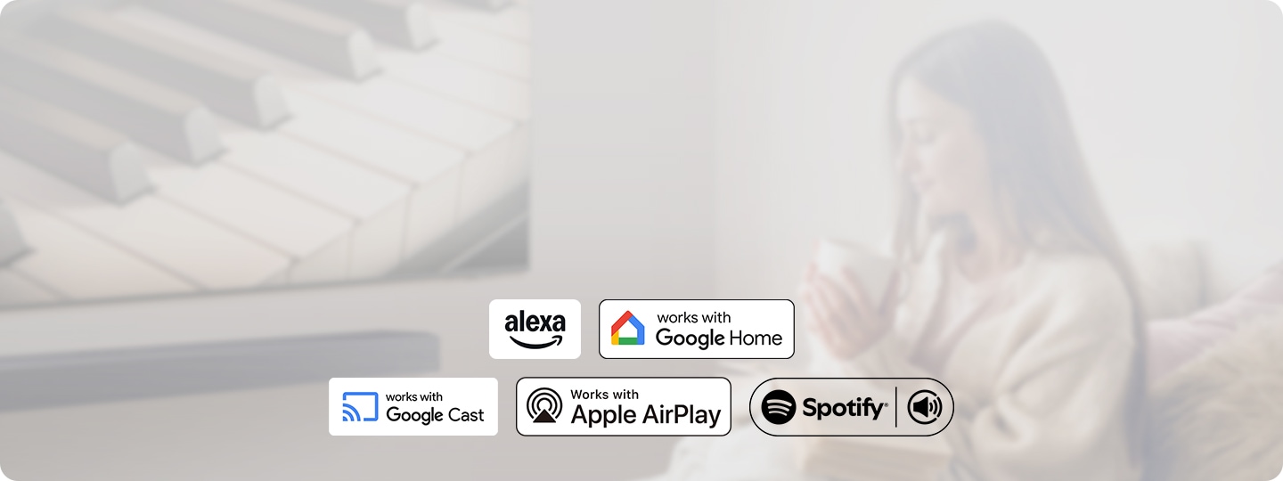 벽에 TV가 설치되어 있으며, TV 하단에는 사운드바가 놓여 있습니다. TV 앞에는 머그컵을 들고 있는 여성이 앉아 있습니다. 이미지 하단에는 알렉사, 구글 홈, 구글 캐스트, 애플 에어플레이, 스포티파이 아이콘이 보입니다