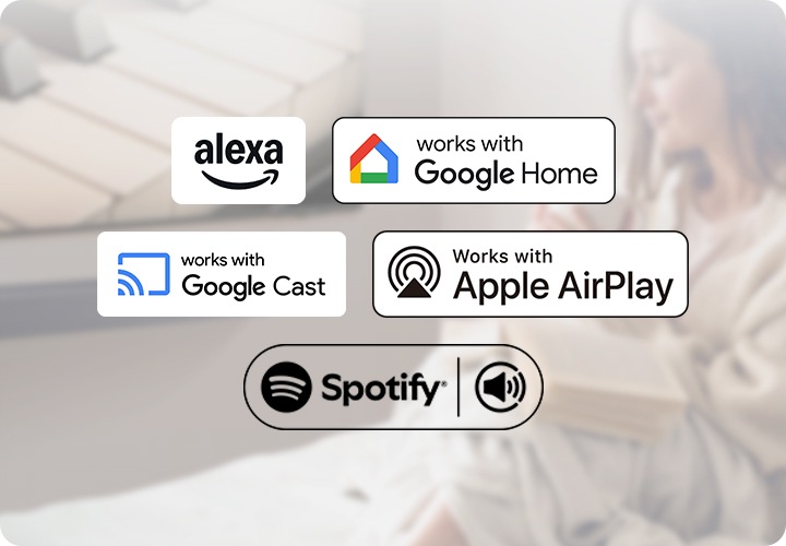 벽에 TV가 설치되어 있으며, TV 하단에는 사운드바가 놓여 있습니다. TV 앞에는 머그컵을 들고 있는 여성이 앉아 있습니다. 이미지 하단에는 알렉사, 구글 홈, 구글 캐스트, 애플 에어플레이, 스포티파이 아이콘이 보입니다