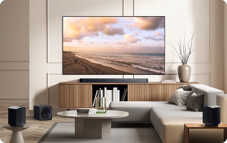 거실 벽에 TV가 설치되어 있으며, TV 아래에는 사운드바가 놓인 장식장이 있습니다. 장식장 오른쪽 상단에는 책과 화병이 놓여 있으며, 장식장 안에는 책들이 꽂혀 있습니다. 장식장 왼쪽에는 서브우퍼가 놓여 있습니다. TV 앞으로는 테이블과 소파, 작은 협탁이 있습니다. 협탁 위에는 무선 리어 스피커가 놓여 있습니다.