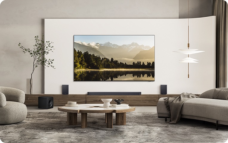 거실 벽에 TV가 설치되어 있으며, TV 아래에는 사운드바와 리어 스피커가 놓인 장식장이 있습니다. 장식장 왼쪽 앞에는 서브우퍼가 놓여 있습니다. TV 앞으로는 테이블과 소파가 놓여 있습니다.