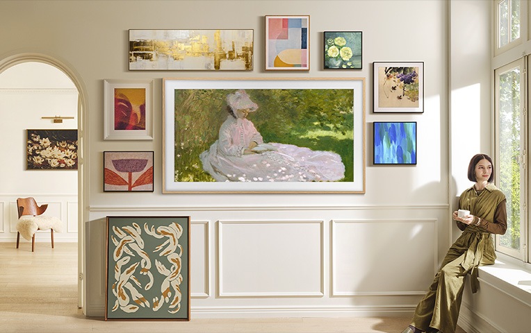 여성이 창틀에 앉아 있습니다. 벽에는 The Frame과 액자들이 걸려 있습니다. TV와 액자에는 명화들이 보여지고 있습니다.