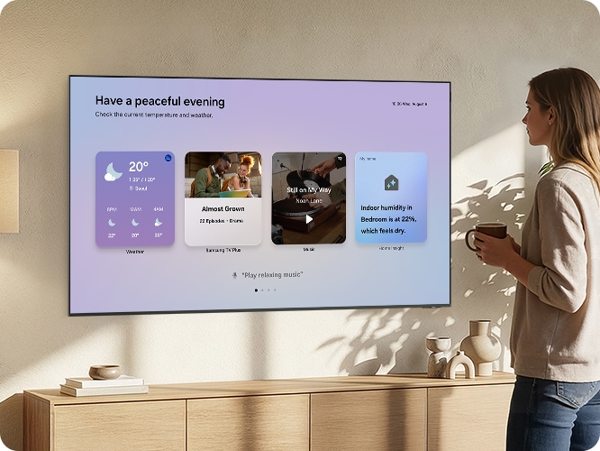 거실 벽에 TV가 설치되어 있으며, TV 화면에는 위젯이 보이고 있습니다. 머그잔을 손에 쥔 여성이 TV를 보고 있습니다.