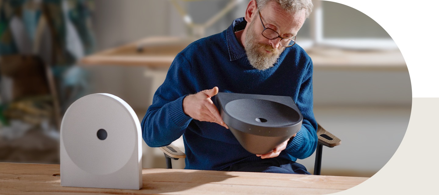 책상 앞 의자에 앉은 디자이너 에르완 부훌렉이 블랙 색상의 스피커를 들어서 보고 있습니다. 책상 위에는 화이트 색상의 스피커가 놓여 있습니다.