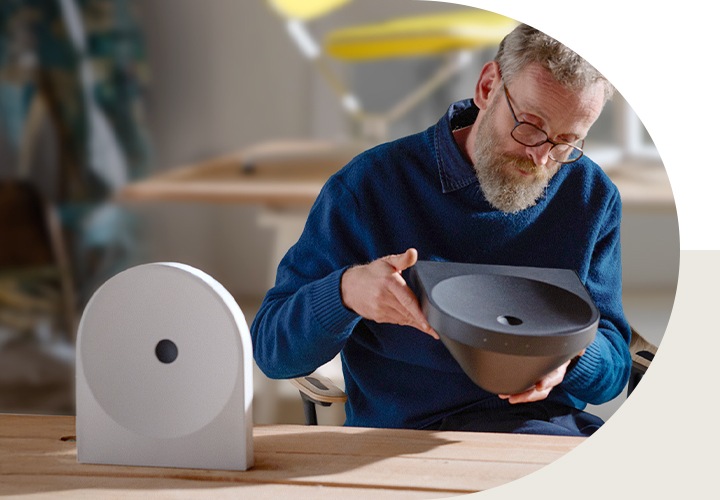 책상 앞 의자에 앉은 디자이너 에르완 부훌렉이 블랙 색상의 스피커를 들어서 보고 있습니다. 책상 위에는 화이트 색상의 스피커가 놓여 있습니다.