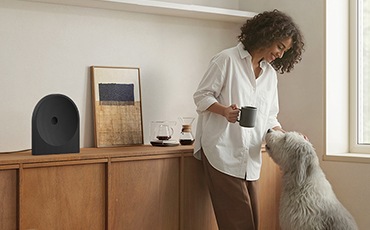 장식장 위에 스피커가 놓여 있습니다. 스피커 오른쪽에는 액자와 장식품이 있습니다. 장식장 앞에는 컵을 들고 있는 여성이 강아지를 보며 웃고 있습니다.