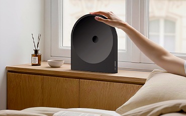장식장 위에 스피커가 놓여 있습니다. 장식장 오른쪽에는 침대가 있으며, 침대에 누워 있는 사람이 스피커를 만지고 있습니다.