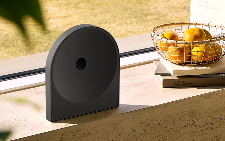 빛이 들어오는 공간에 스피커가 놓여 있습니다. 스피커 오른쪽에는 귤이 담긴 바구니와 책들이 보입니다.