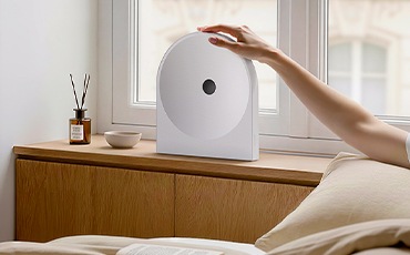 장식장 위에 스피커가 놓여 있습니다. 장식장 오른쪽에는 침대가 있으며, 침대에 누워 있는 사람이 스피커를 만지고 있습니다.
