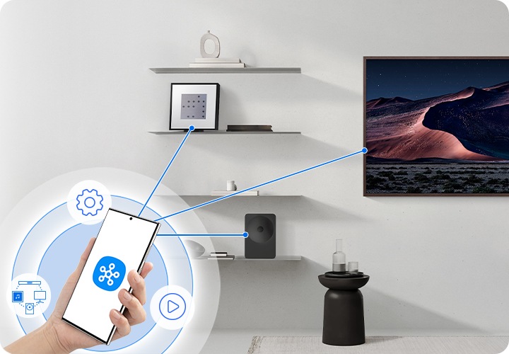 왼쪽 하단에 휴대폰을 쥐고 있는 손이 있습니다. 휴대폰 화면에는 스마트싱스 앱 아이콘이 있으며, 휴대폰에서부터 스피커와 TV쪽으로 선이 연결되어 있습니다.