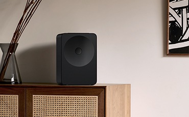 장식장 위에 스피커가 놓여 있습니다. 장식장 왼쪽에는 화병이 놓여 있고, 오른쪽 벽에는 액자가 걸려 있습니다.