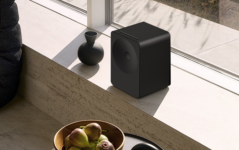 빛이 들어오는 공간에 스피커가 놓여 있습니다. 스피커 왼쪽에는 화병이 있습니다. 스피커 앞에는 무화과가 담긴 바구니가 있습니다.