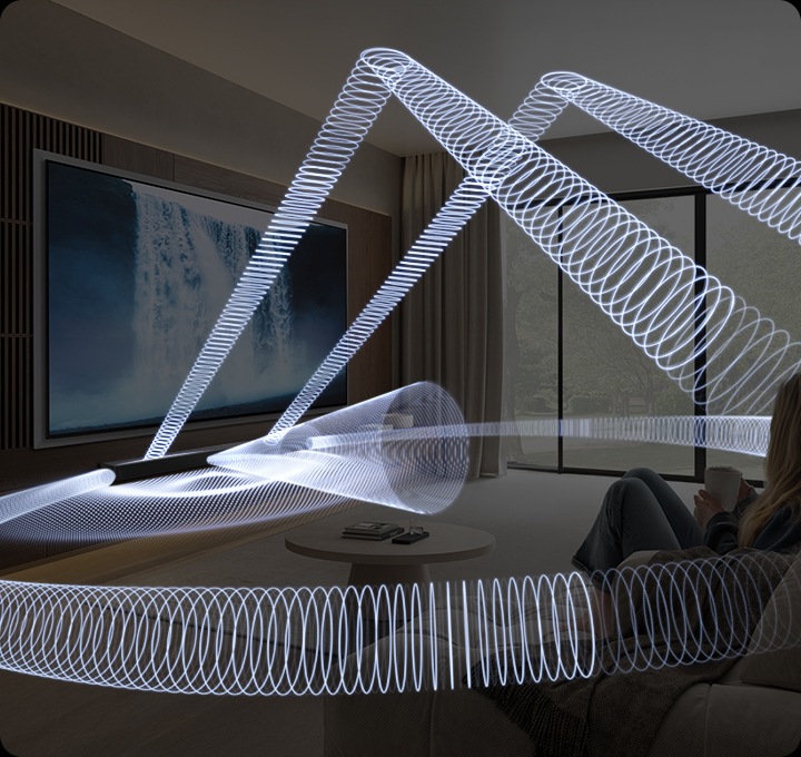 소파에 앉아있는 여성이 거실 벽에 설치된 TV를 보고 있습니다. TV 화면에는 폭포의 모습이 보입니다. TV 하단에는 사운드바가 설치되어 있으며, 사운드바에서 음향효과가 나옵니다.