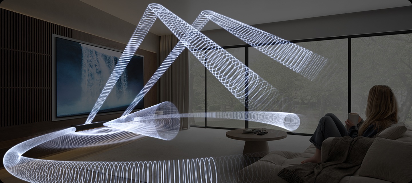 소파에 앉아있는 여성이 거실 벽에 설치된 TV를 보고 있습니다. TV 화면에는 폭포의 모습이 보입니다. TV 하단에는 사운드바가 설치되어 있으며, 사운드바에서 음향효과가 나옵니다.