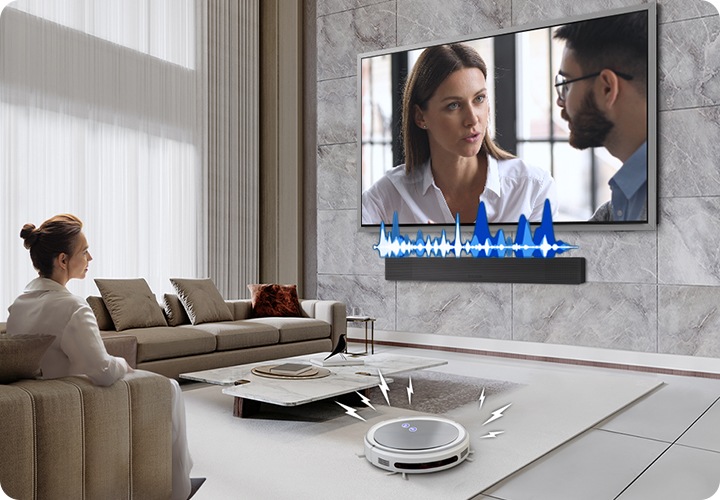 거실에서 여성이 TV를 보고 있습니다. TV 화면에는 드라마에 한 장면이 나오고 있습니다. TV 하단에는 사운드바가 설치되어 있고 TV와 사운드바 사이에는 음향효과가 있습니다. 거실 바닥에는 무선 로봇 청소기가 움직이고 있습니다.
