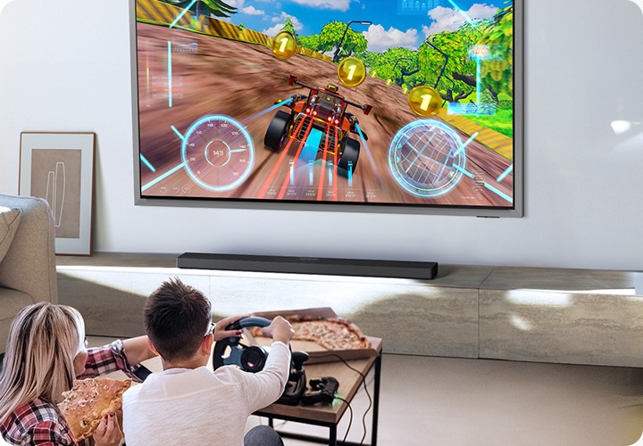 벽에 TV가 설치되어 있습니다. TV 아래에는 사운드바가 놓인 장식장이 있습니다. TV 앞에는 남성과 여성이 거실에 앉아서 게임을 즐기고 있습니다.
