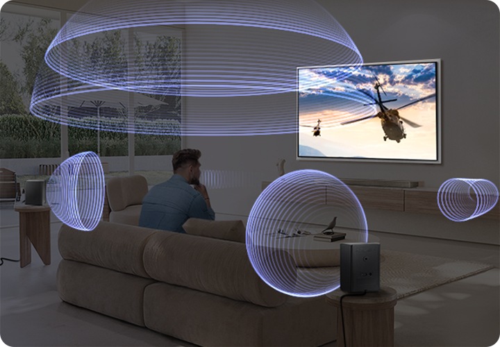 거실 벽에 TV가 설치되어 있습니다 TV 하단에는 장식장이 있으며, 그 위 사운드바가 놓여 있습니다. TV 화면에는 헬리콥터가 하늘을 날고 있는 모습이 보입니다. TV 앞에는 소파가 있으며, 소파에 남성이 앉아 있습니다. 소파 뒤로는 협탁이 있으며 그 위에 리어 스피커가 설치되어 있습니다. 사운드바와 리어 스피커에서 음향효과가 나오고 있습니다.