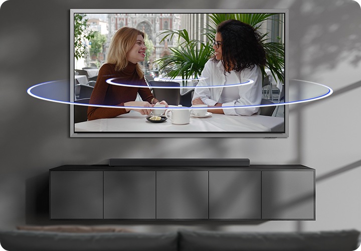 벽에 TV가 설치되어 있으며, TV 아래에는 사운드바가 놓인 장식장이 있습니다. TV 화면에는 여성 두명이 티타임을 가지고 있습니다. TV 화면에서 음향효과가 나오고 있습니다.