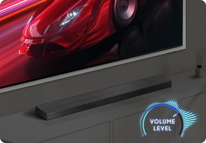 벽에 TV가 설치되어 있으며, TV 아래에는 사운드바가 놓인 장식장이 있습니다. TV 화면에는 붉은색 자동차가 송출됩니다. 이미지 오른쪽 하단에는 볼륨 레벨이 표시되어 있습니다.