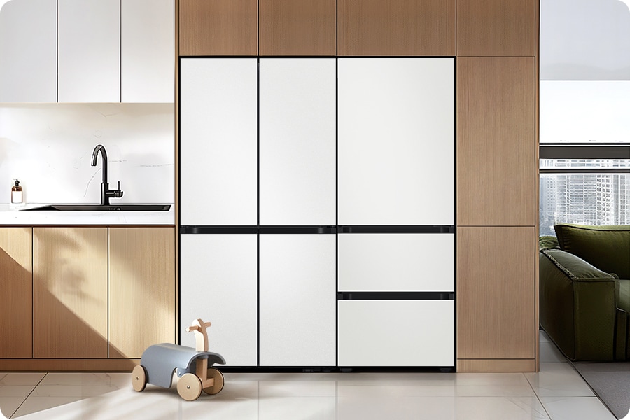 3인 가구. 우드톤 냉장고장에 냉장고 4도어와 김치플러스 3도어가 페어로 설치되어 있습니다. 냉장고 앞에는 아기 장난감이 놓여있습니다. 냉장고장 좌측에는 하얀 상부장과 우드톤 하부장 위에 수전이 설치되어 있습니다. 냉장고장 우측에는 녹색 쇼파가 보이고 뒤로 통창이 있는 연출된 이미지 입니다.