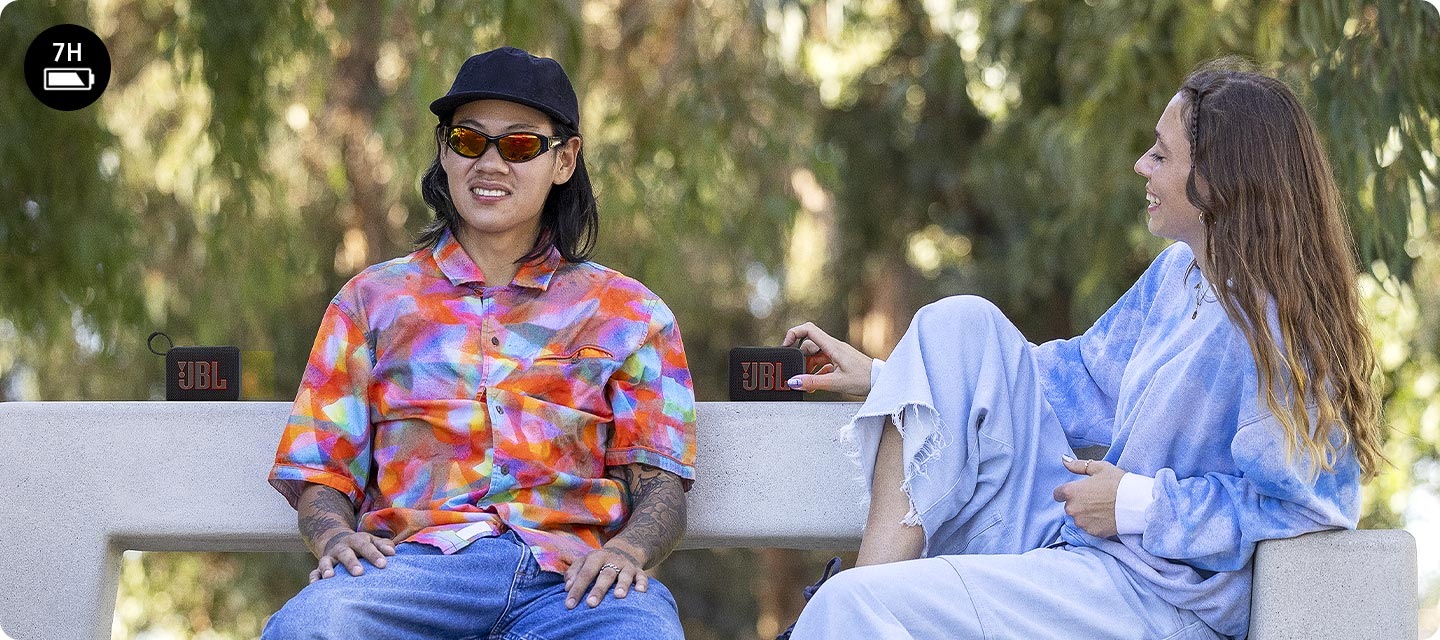 남성과 여성이 벤치에 앉아 있습니다. 왼쪽에 앉아 있는 남성의 뒤로 스피커 한 대가 놓여 있으며, 오른쪽에 앉아 있는 여성의 오른손에 스피커 한 대가 있습니다.