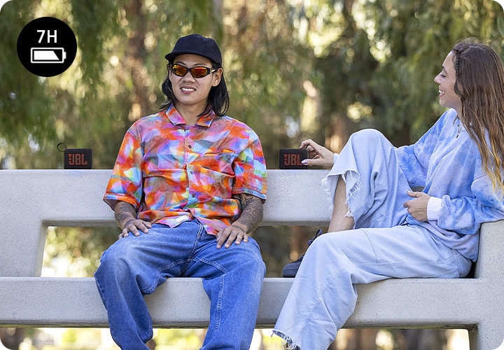 남성과 여성이 벤치에 앉아 있습니다. 왼쪽에 앉아 있는 남성의 뒤로 스피커 한 대가 놓여 있으며, 오른쪽에 앉아 있는 여성의 오른손에 스피커 한 대가 있습니다.