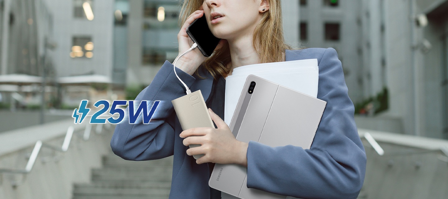 정장 차림의 여성이 갤럭시 S22로 통화 중입니다. 기기는 USB-C 케이블로 배터리팩에 연결되어 손 안에서 충전되고 있습니다. 동시에 갤럭시 탭 S8+와 몇 장의 서류도 들고 있습니다. 왼쪽에는 25W가 표시되어 있습니다.