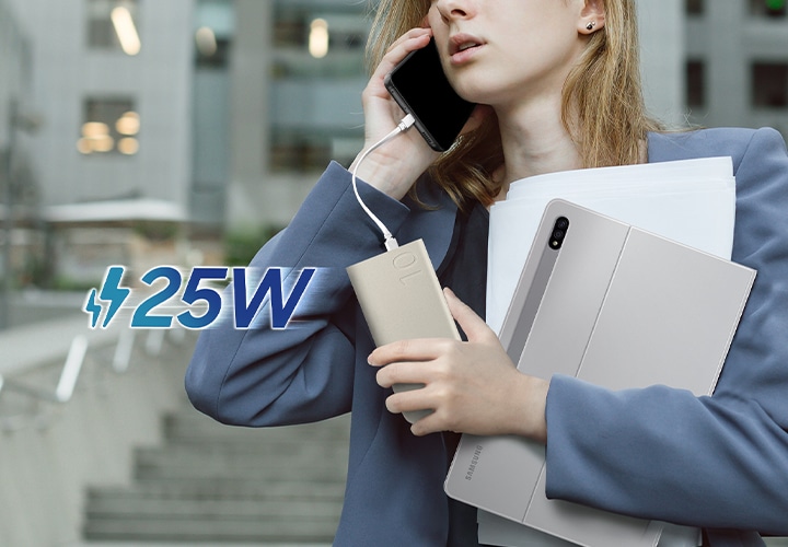 정장 차림의 여성이 갤럭시 S22로 통화 중입니다. 기기는 USB-C 케이블로 배터리팩에 연결되어 손 안에서 충전되고 있습니다. 동시에 갤럭시 탭 S8+와 몇 장의 서류도 들고 있습니다. 왼쪽에는 25W가 표시되어 있습니다.