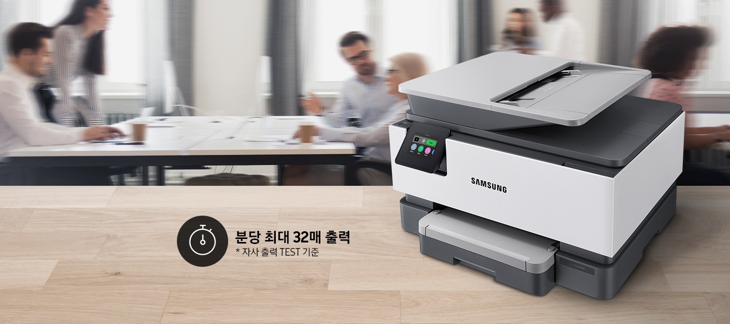 우드 패턴 테이블 위에 잉크젯 복합기가 놓여있고, 여러 사람이 있는 사무실이 뒤 배경으로 있습니다.