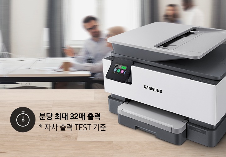 우드 패턴 테이블 위에 잉크젯 복합기가 놓여있고, 여러 사람이 있는 사무실이 뒤 배경으로 있습니다.