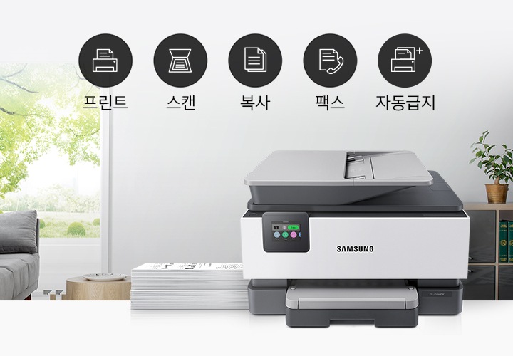 창 밖으로 나무가 보이는 배경 앞에 잉크젯 복합기와 그 옆에 프린트물이 쌓여있습니다. 왼쪽 공간에는 프린트, 스캔, 복사, 팩스, 자동급지 아이콘이 있습니다.