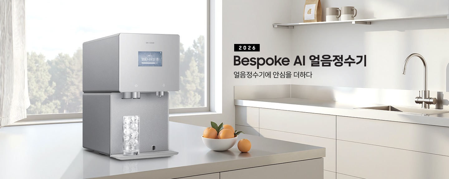 2026 Bespoke AI 얼음정수기. 얼음정수기에 안심을 더하다.  회색 테이블 위에 얼음정수기 사틴 그레이지 컬러가 설치되어 있는 측면 이미지 입니다. 정수기 얼음 토출구 아래에는 얼음이 가득 든 물컵이 놓여있습니다. 정수기 우측에는 오렌지가 가득 담긴 그릇이 놓여있습니다.