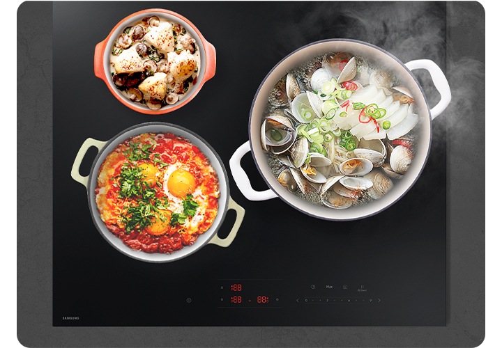 진한 회색 상판의 대리석을 배경으로 이미지 중간 부분에는 인덕션이 설치되어 있고 인덕션 위에는 각각 크기가 다른 냄비 3개에서 음식을 조리 중인 모습이 있습니다. 인덕션 오른쪽 부분에는 그릇 안에 나무 국자가 놓여 있습니다.